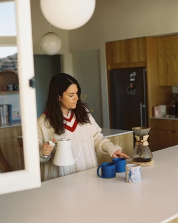 Woman preparing pour-over coffee at a white kitchen counter with mugs, a kettle, and kitchen cabinets visible in soft, natural light.