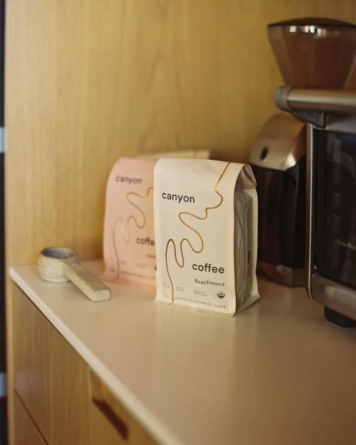 Close-up of two Canyon Coffee bags, one pink and one white, alongside a ceramic spoon on a counter with a coffee grinder in the background.
