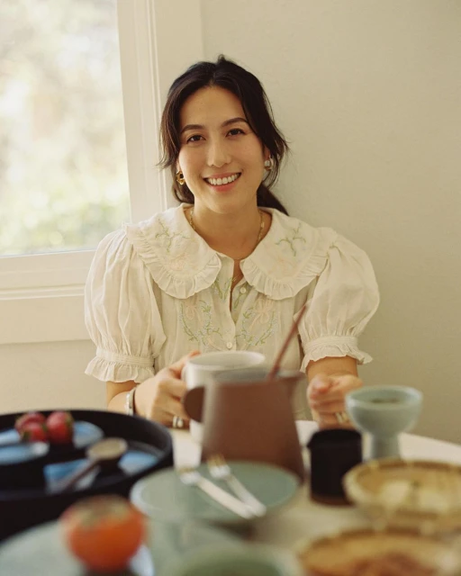 Adult female smiles near a table with ceramic dishware, a brown pitcher, and tomatoes in a bright indoor dining setting.