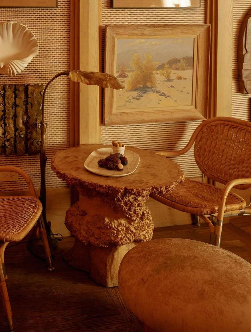 Interior shot of a wicker chair and wooden table with food, alongside a lamp and decorative wall art, evoking a cozy and rustic ambiance.