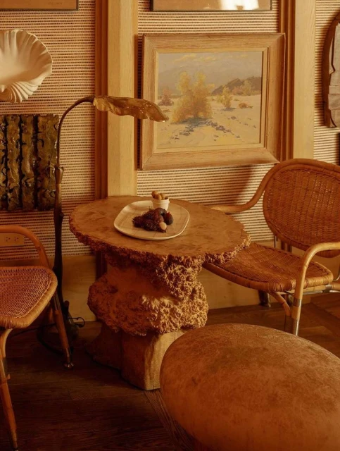 Interior shot of a wicker chair and wooden table with food, alongside a lamp and decorative wall art, evoking a cozy and rustic ambiance.