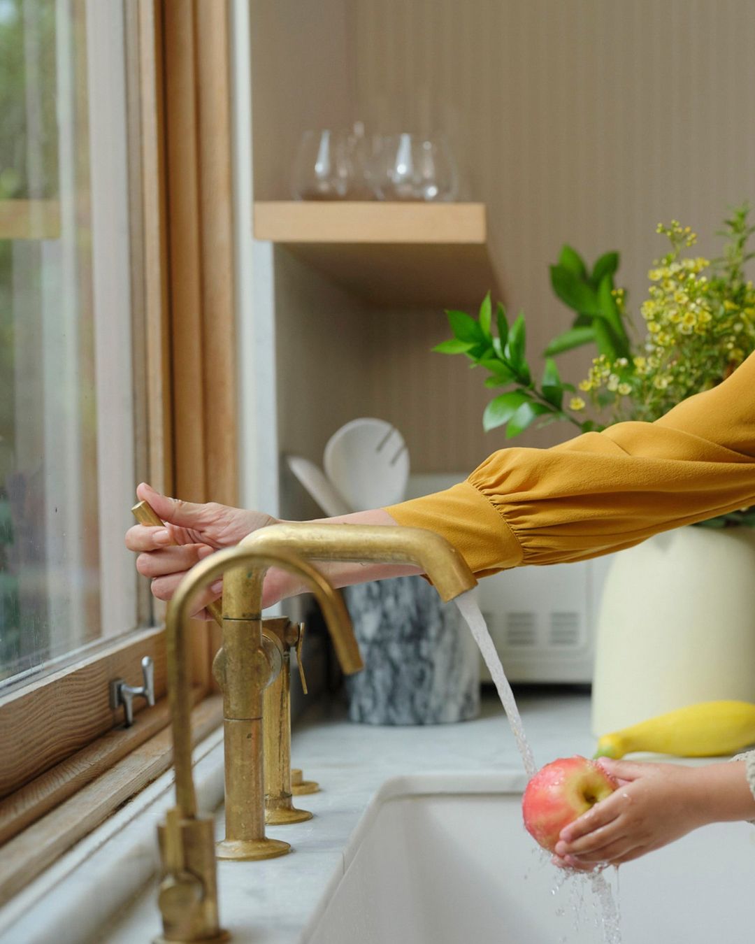 Child washes an apple under a gold faucet in a bright kitchen with a window, while an adult hand adjusts the water.