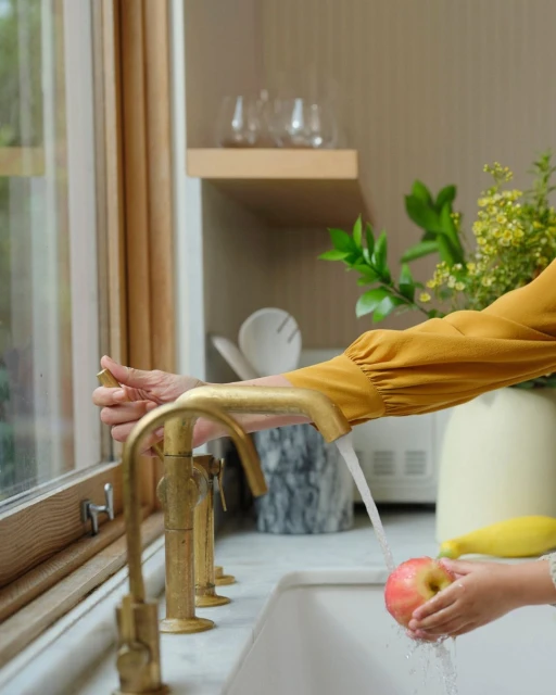 Child washes an apple under a gold faucet in a bright kitchen with a window, while an adult hand adjusts the water.