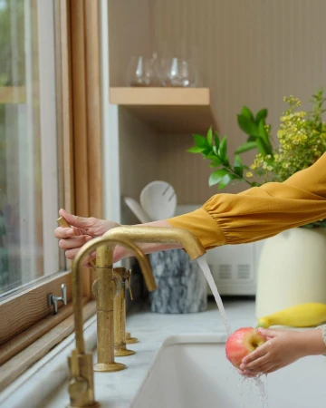Child washes an apple under a gold faucet in a bright kitchen with a window, while an adult hand adjusts the water.