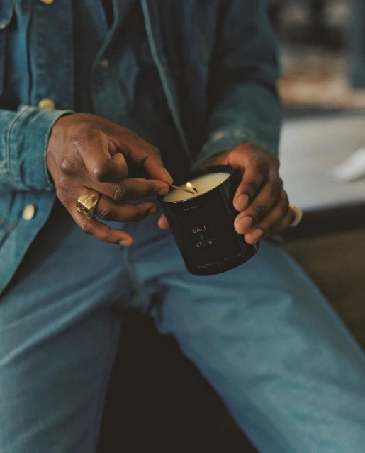 An adult male in denim lights a candle with a match, revealing the candle's 'Salt & Stone' label, in a warmly lit, indoor setting.
