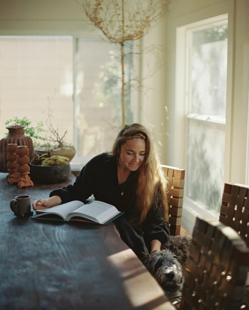 Woman reads book with dog beside her on long, dark wood table near window and plants in bright, naturally lit room.