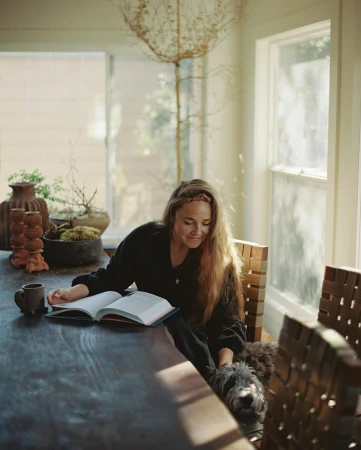 Woman reads book with dog beside her on long, dark wood table near window and plants in bright, naturally lit room.