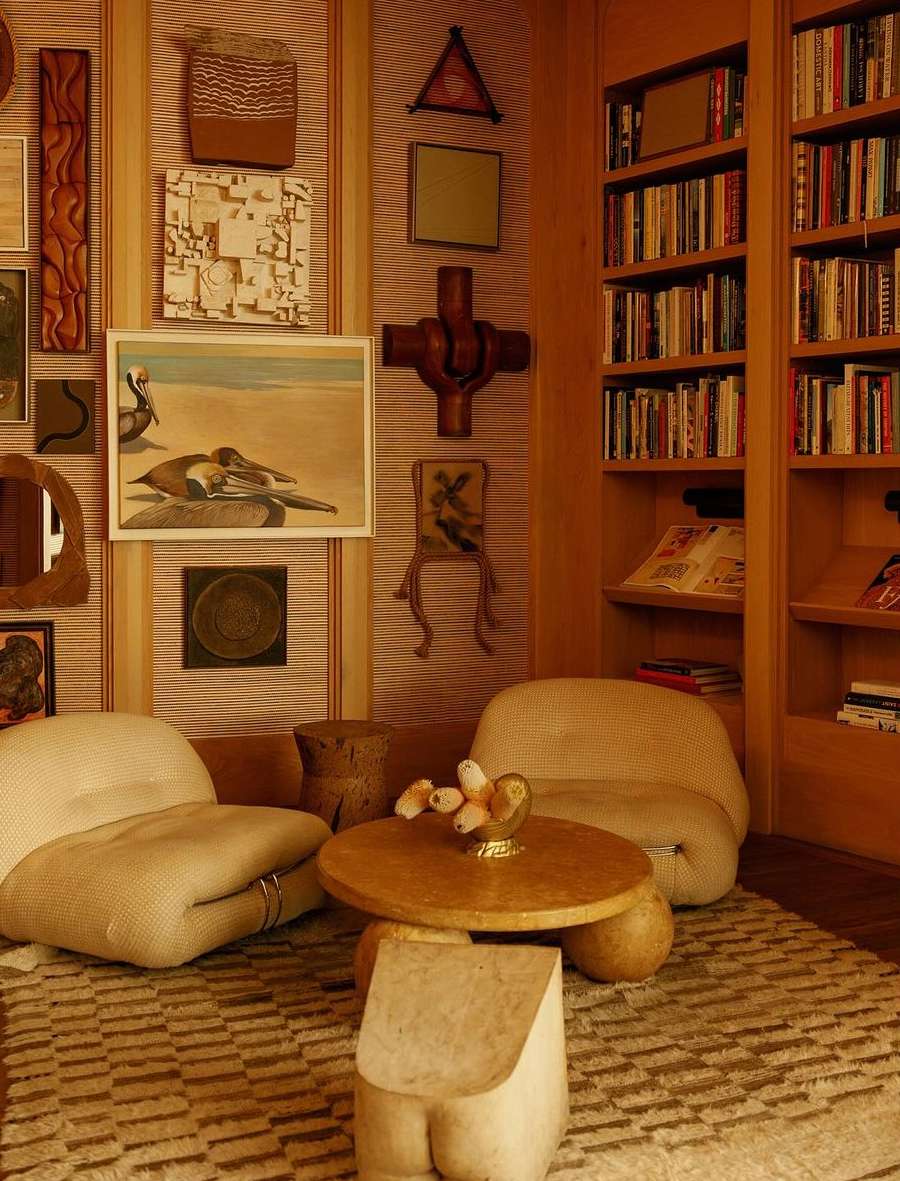 Warm-toned interior with two armchairs, a wooden coffee table, and built-in bookshelves filled with books and decorative art pieces on the wall.