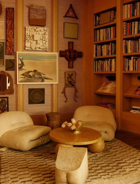 Warm-toned interior with two armchairs, a wooden coffee table, and built-in bookshelves filled with books and decorative art pieces on the wall.