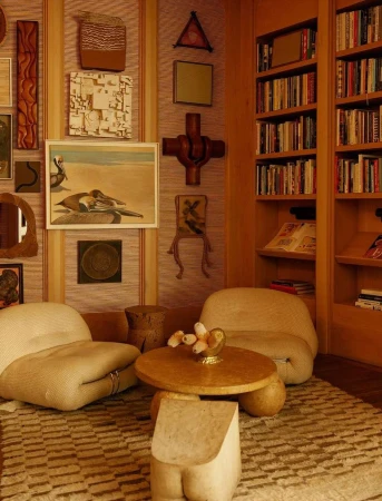 Warm-toned interior with two armchairs, a wooden coffee table, and built-in bookshelves filled with books and decorative art pieces on the wall.