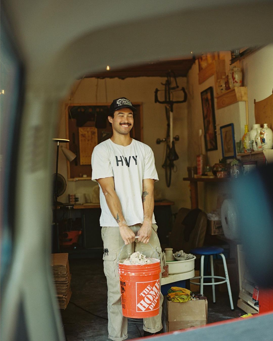 Young man in white t-shirt holds an orange bucket filled with white clay in his pottery studio, looking away from the camera and smiling.