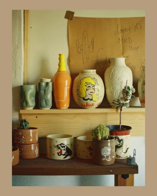 Still life shot of earthenware pottery and potted plants displayed on a wooden shelf against a neutral wall with hand-drawn cactus art.