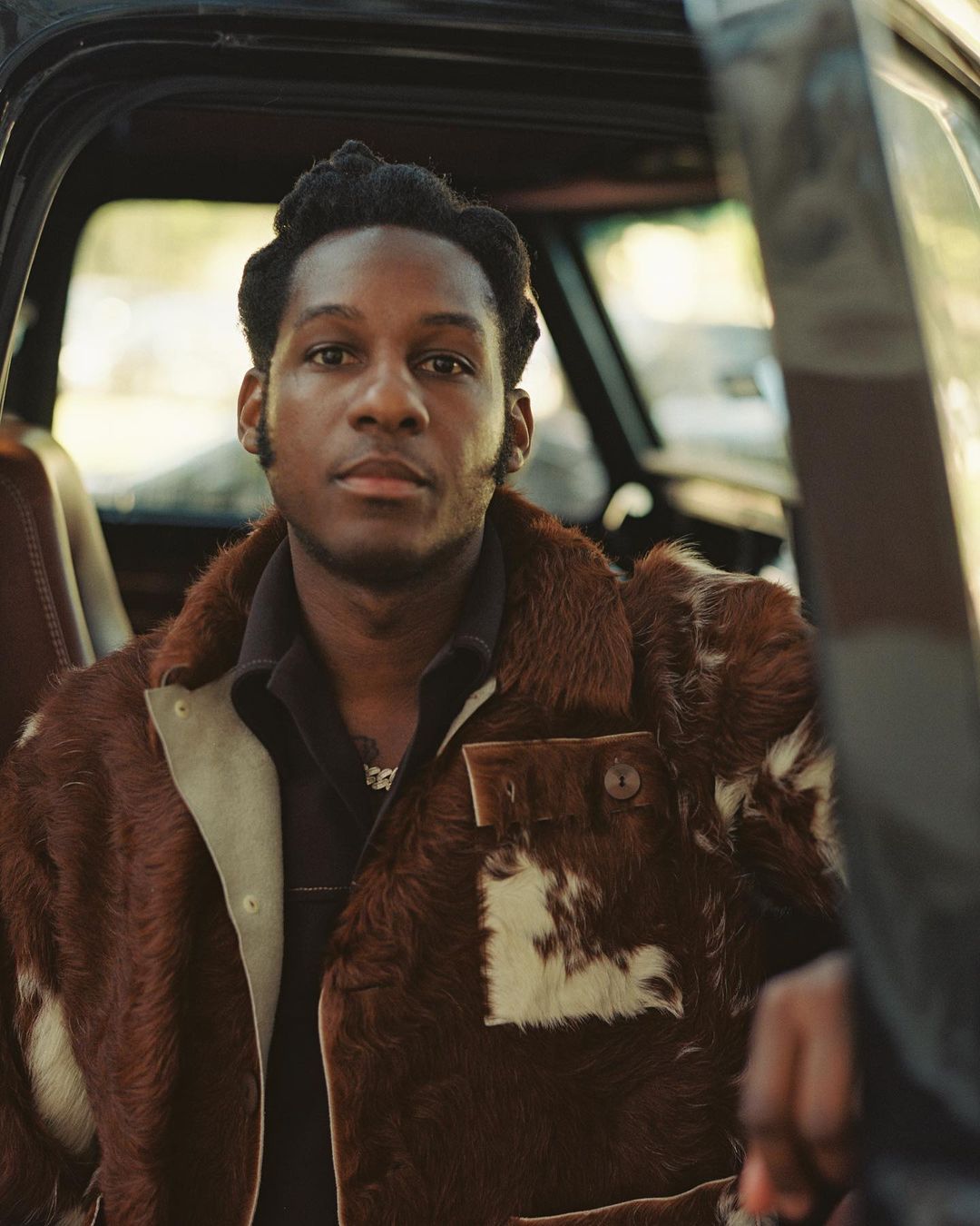 Portrait of an African-American adult male seated in a black truck wearing a brown and white patterned fur coat with a black collared shirt.
