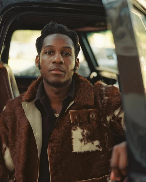 Portrait of an African-American adult male seated in a black truck wearing a brown and white patterned fur coat with a black collared shirt.