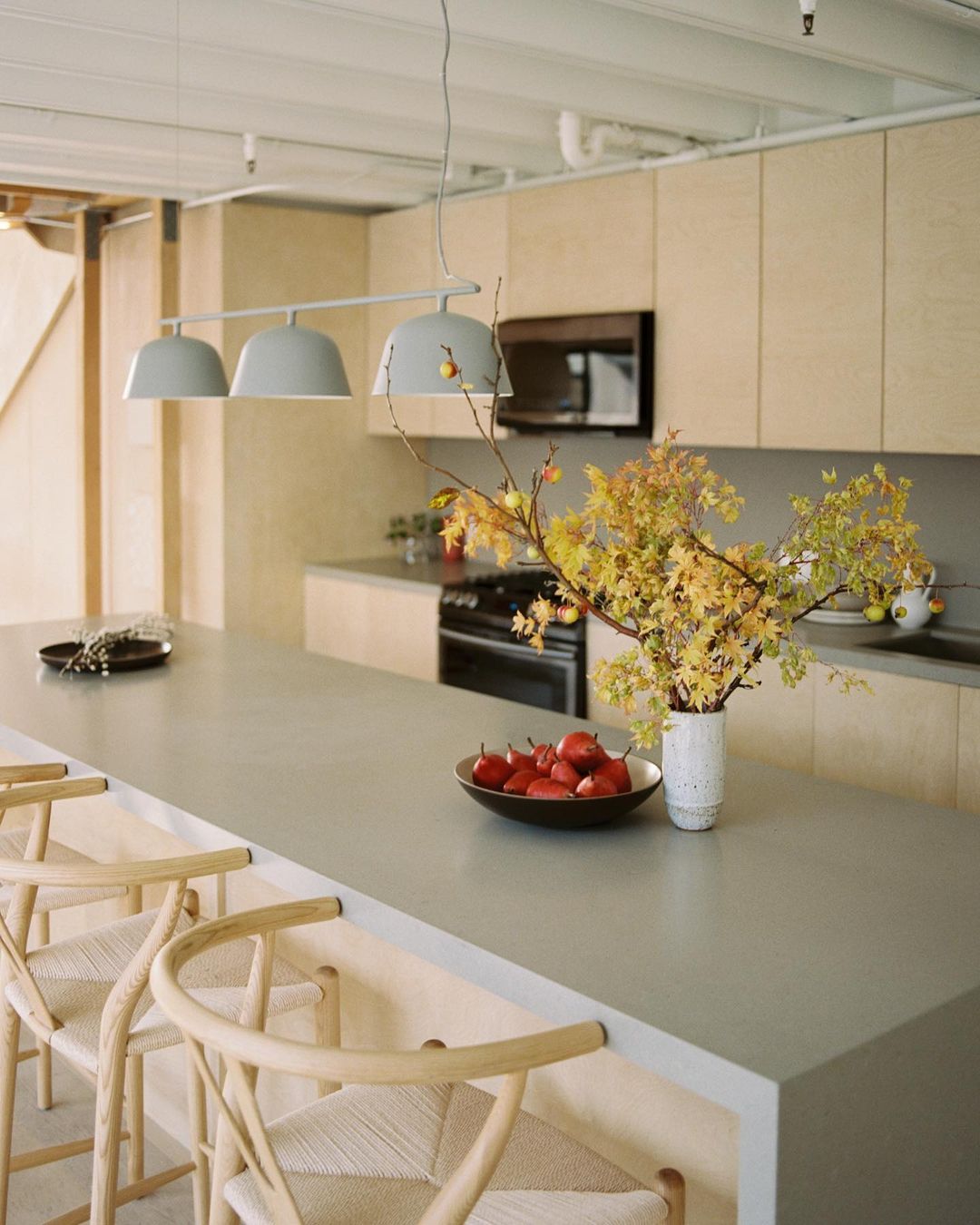 Contemporary kitchen interior featuring a gray countertop island with stools, pendant lighting, fruit bowl, vase with branches and cabinets.