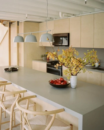 Contemporary kitchen interior featuring a gray countertop island with stools, pendant lighting, fruit bowl, vase with branches and cabinets.