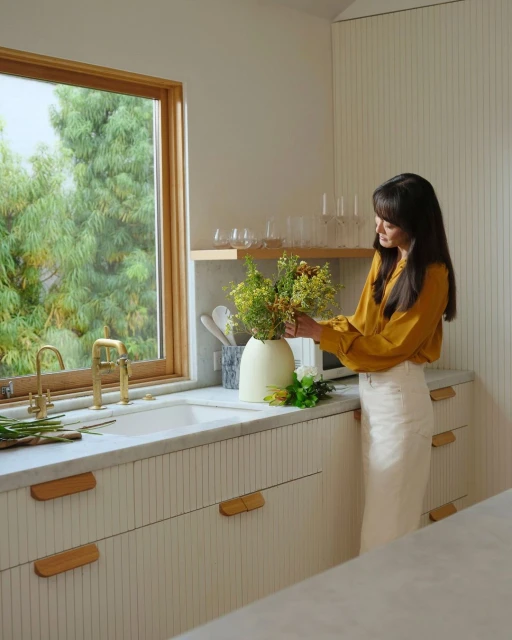 Woman arranging vase of flowers in bright, modern kitchen with white cabinetry, wood accents, and natural light streaming through window.