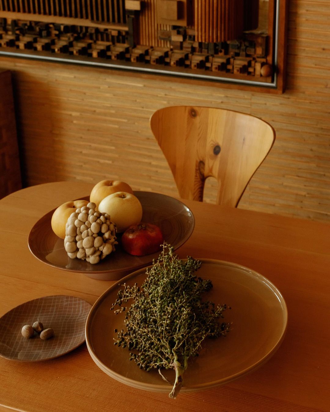 Still life on a wooden table featuring apples, decorative balls, and a sprig of greenery, under warm indoor lighting, with an abstract wood sculpture in the background.