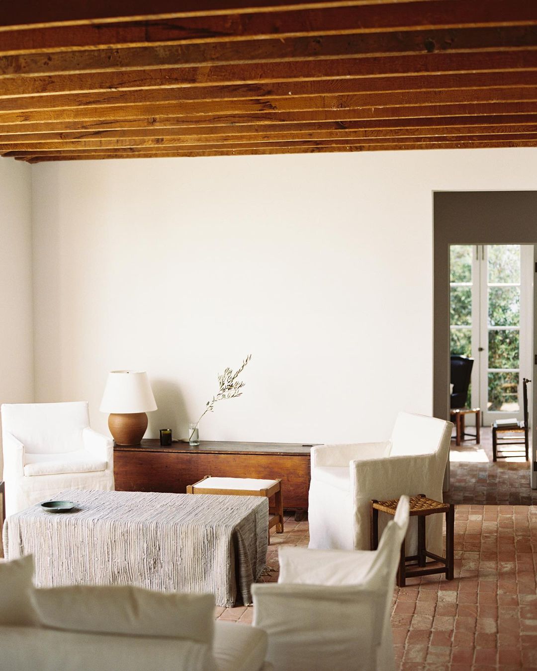 Living room interior with white armchairs, wooden coffee tables, and brick flooring under a wooden ceiling.