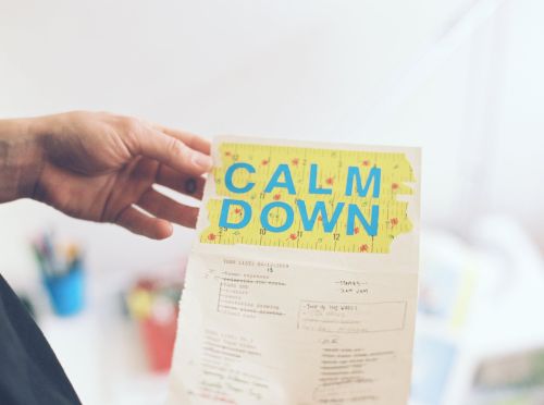 Close-up of a hand holding up a paper with the words 'Calm Down' in blue block letters taped to the top. The background is out of focus.