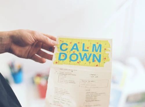 Close-up of a hand holding up a paper with the words 'Calm Down' in blue block letters taped to the top. The background is out of focus.
