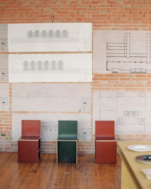 Three colorful chairs sit in a room with exposed brick walls, and architectural drawings hung on the wall, showcasing interior design elements.
