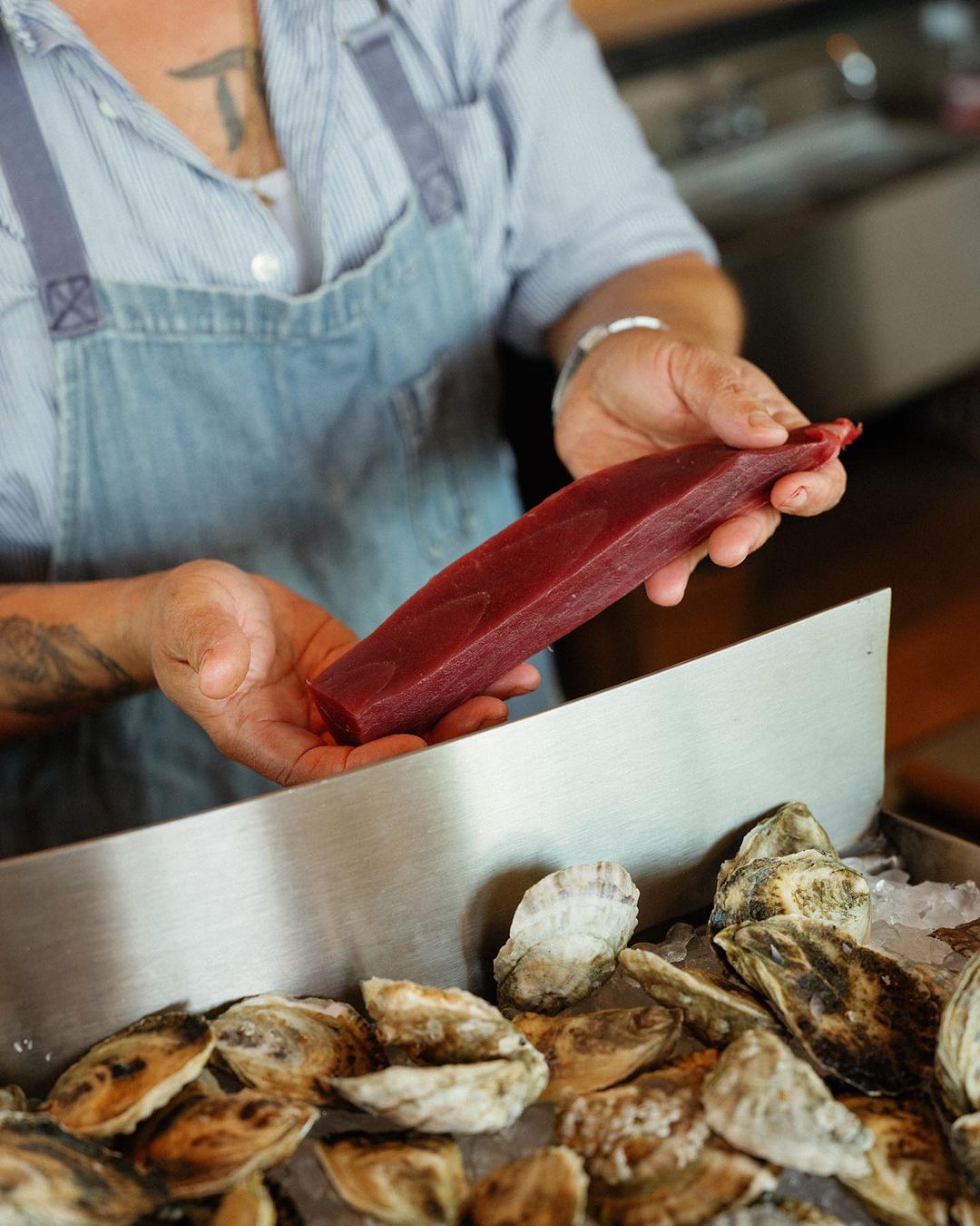 Chef holding a raw tuna steak over a tray of oysters on ice, wearing a denim apron, in a restaurant setting with natural light.