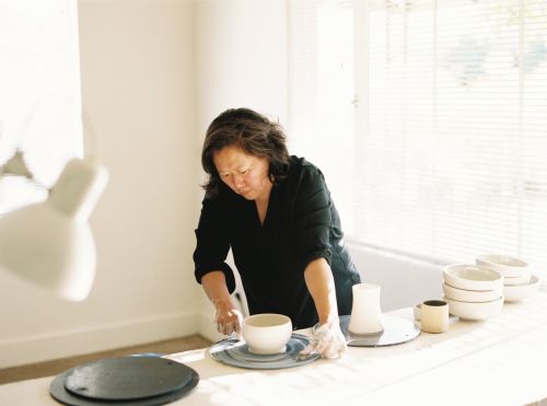 Artist crafting a ceramic bowl on a table in a bright studio, showcasing pottery and craftsmanship in a well-lit setting with neutral tones.
