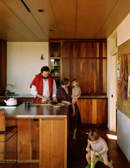 Family in a modern kitchen with wood cabinets, stainless steel countertops, and a kitchen island, one adult is cutting vegetables.