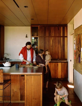 Family in a modern kitchen with wood cabinets, stainless steel countertops, and a kitchen island, one adult is cutting vegetables.