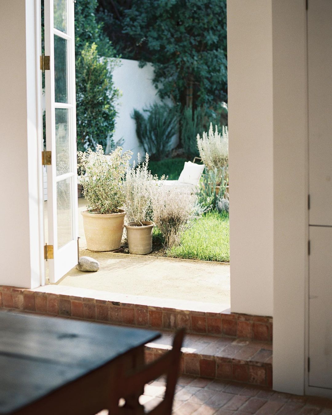 Open French doors lead from an indoor room to an outdoor patio and garden with plants, lawn, and white lounge chair in natural light.