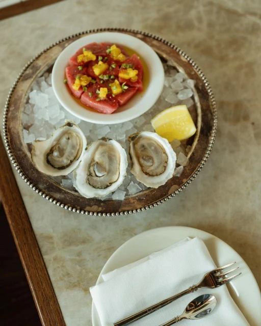 Overhead shot of three oysters on crushed ice with lemon, accompanied by a dish of diced fish with yellow toppings on a silver tray.
