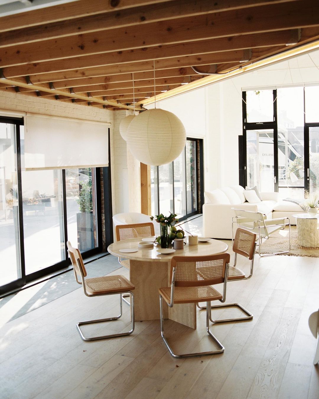 Bright, naturally lit dining area features a round table with four chairs, pendant lights, and large windows with white blinds. Interior design.