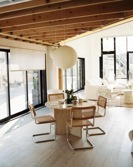 Bright, naturally lit dining area features a round table with four chairs, pendant lights, and large windows with white blinds. Interior design.