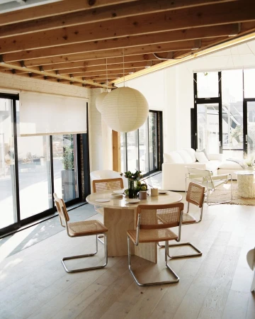 Bright, naturally lit dining area features a round table with four chairs, pendant lights, and large windows with white blinds. Interior design.