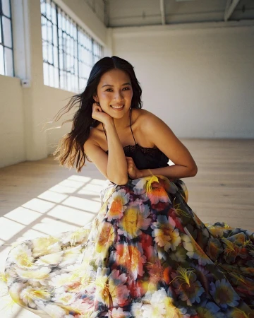 Smiling woman in black top and floral skirt seated on wood floor by large window