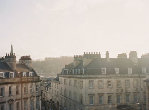 Cityscape view of historic stone buildings in Bath, England, under soft light. Architectural details and distant trees visible.