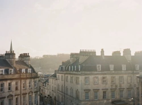 Cityscape view of historic stone buildings in Bath, England, under soft light. Architectural details and distant trees visible.