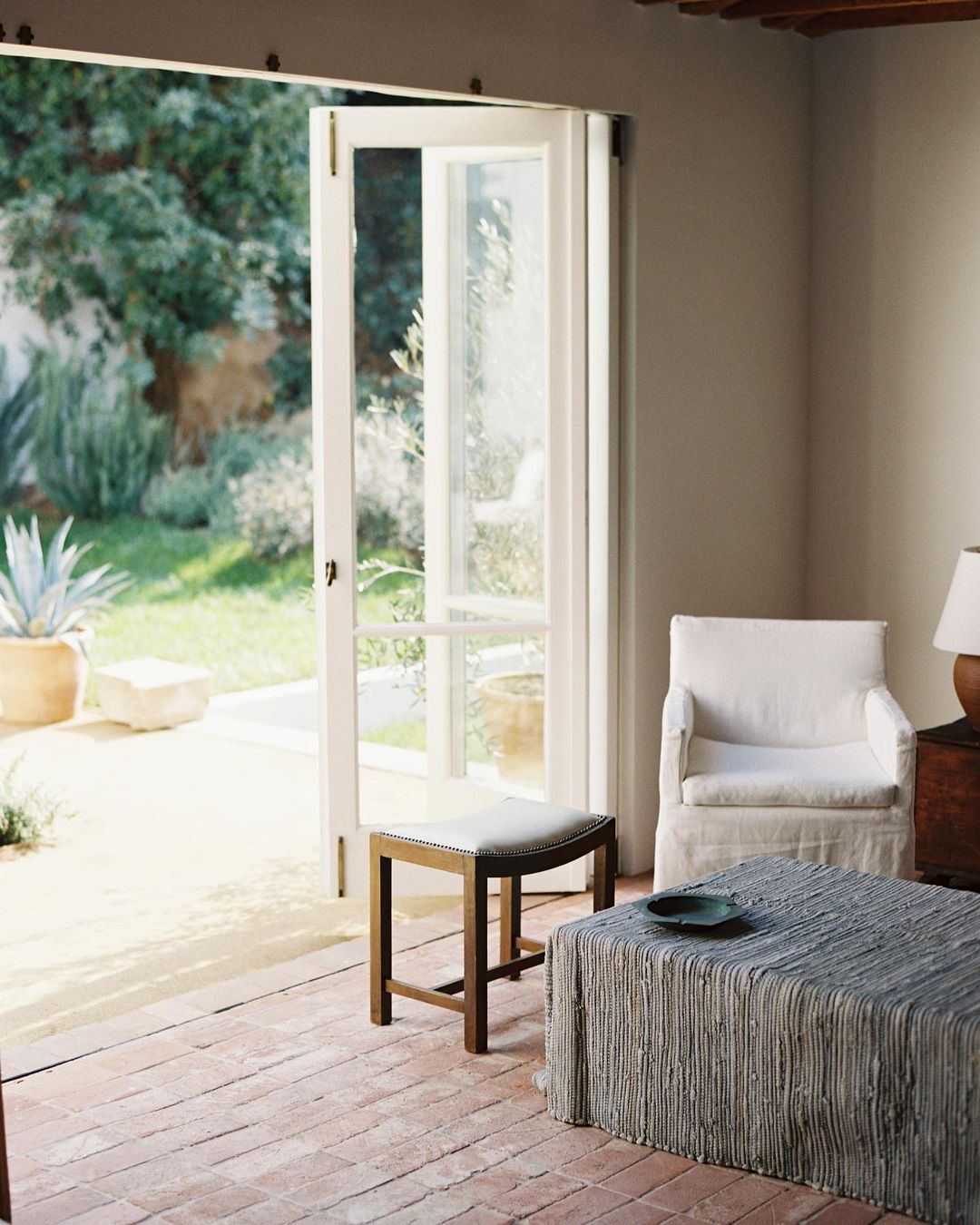 Living room interior with brick floor, white upholstered furniture, an open door to an outdoor garden with potted plants.