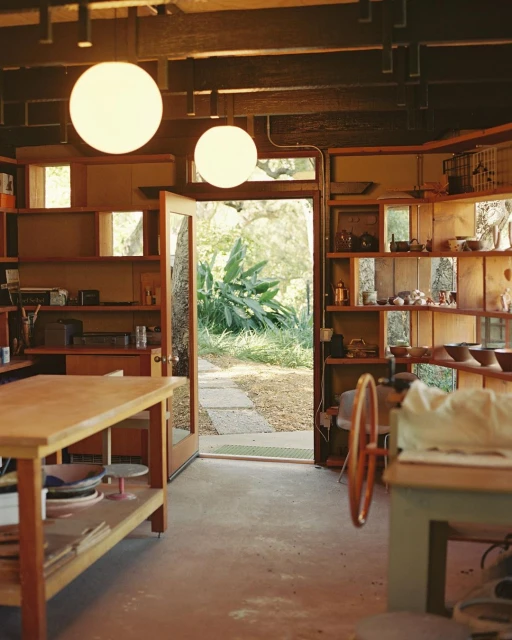 A light-filled pottery studio featuring open shelving filled with ceramic bowls and a potting wheel, viewed from an indoor setting.