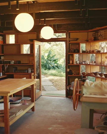 A light-filled pottery studio featuring open shelving filled with ceramic bowls and a potting wheel, viewed from an indoor setting.