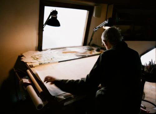 Elderly man drawing at a table illuminated by lamps in a dimly lit room, focused on creative work and architectural design.