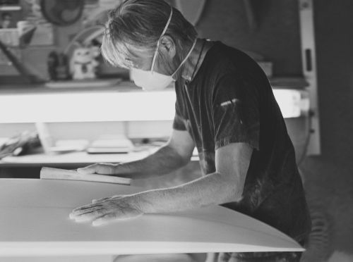 Black and white photo of a man wearing a mask and sanding a surfboard, focused on craftsmanship and surfboard creation indoors.