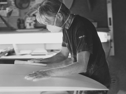 Black and white photo of a man wearing a mask and sanding a surfboard, focused on craftsmanship and surfboard creation indoors.