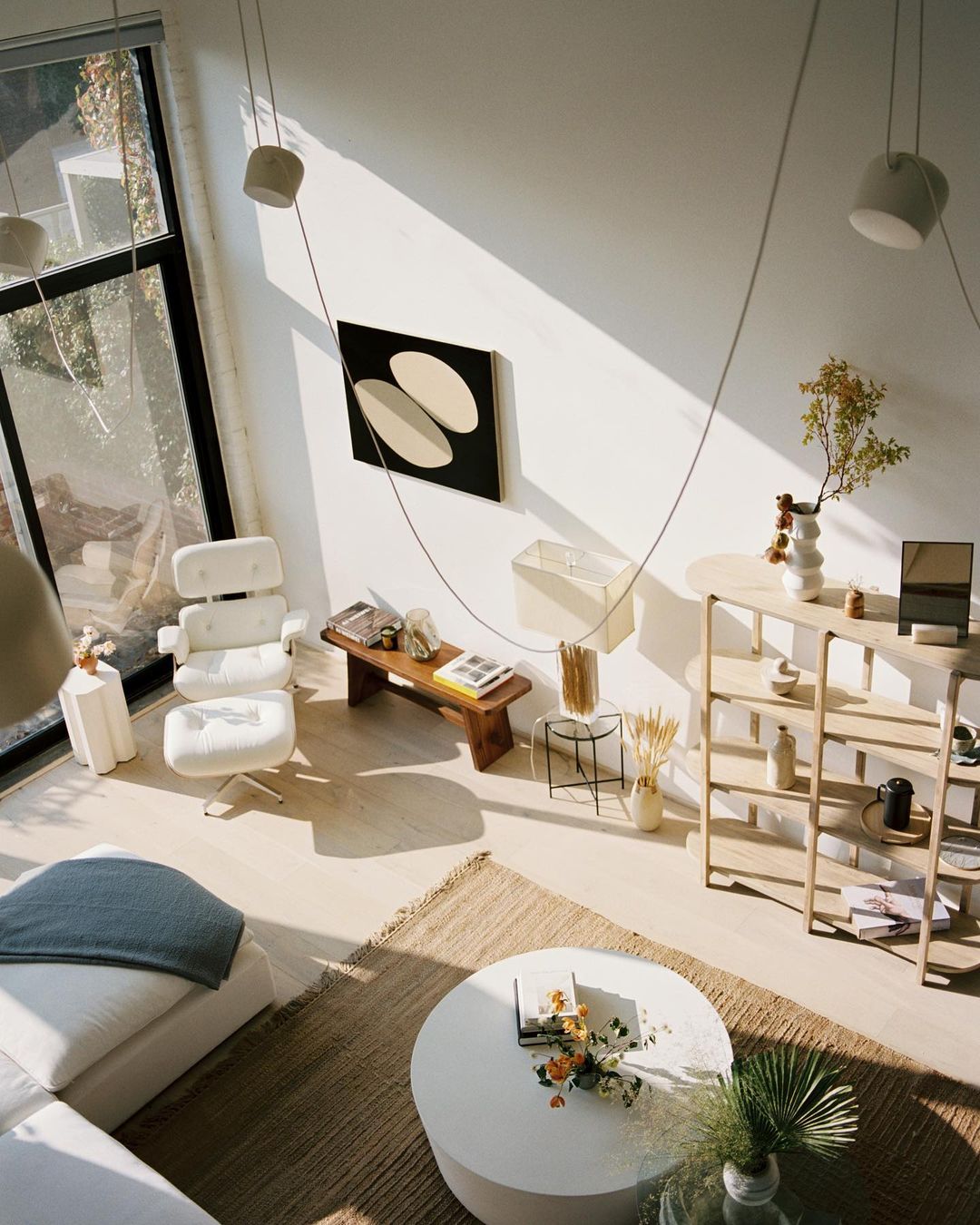 Living room interior with white modern furniture, area rug, abstract art, open-plan design, and natural light from a large window.