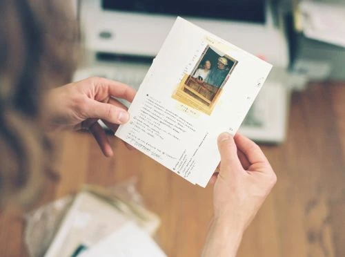 Person holds a paper with a photo of a couple. The paper has handwritten text. A desktop computer is visible in the background.