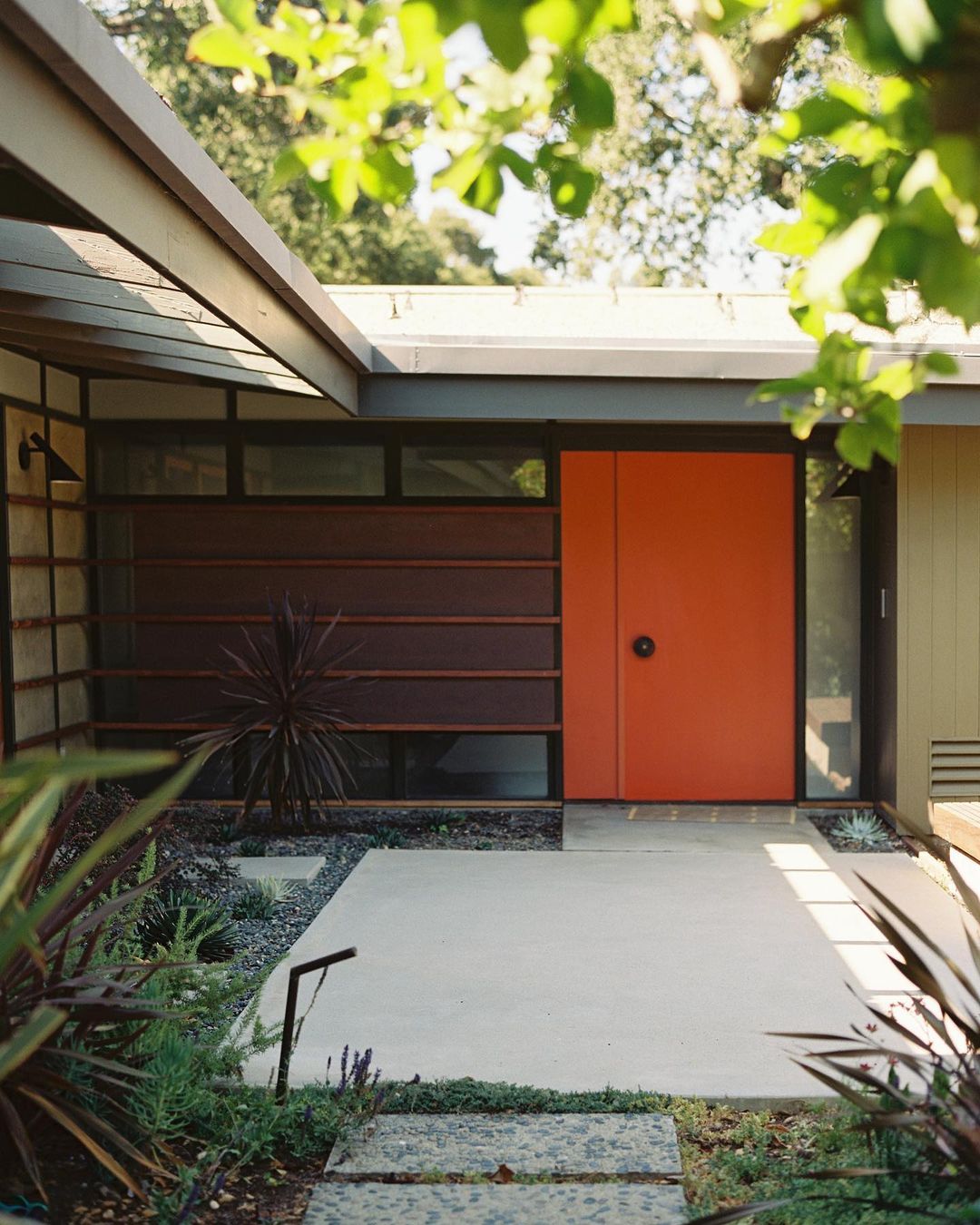 Exterior view of a modern house featuring a bright orange front door, concrete walkway, and garden with green plants and trees.