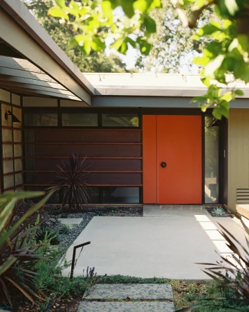 Exterior view of a modern house featuring a bright orange front door, concrete walkway, and garden with green plants and trees.