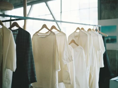 Rack of neutral-colored long-sleeve shirts on wooden hangers, including light cream, white, and a dark blue plaid pattern garment.