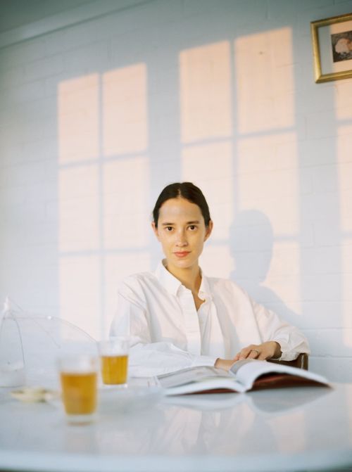 Woman poses indoors at a table with an open book and drinks, window light casting shadows on the white paneled wall behind her.